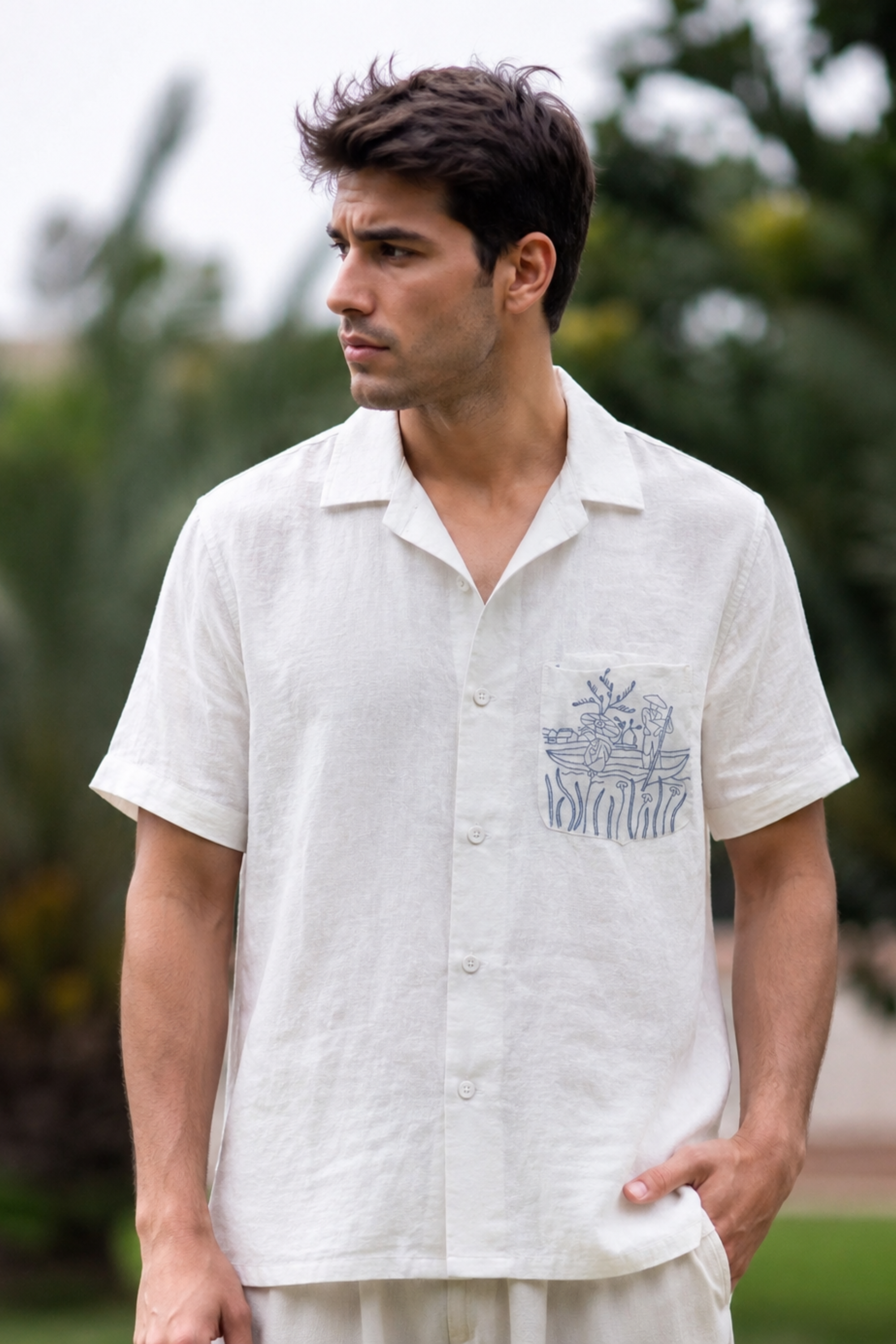 Man wearing a white shirt with a small design, standing outdoors with greenery in the background